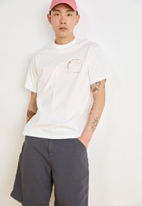 Hvit t-skjorte med korte ermer som har et lite brunt sirkulært grafisk motiv på brystet, sammen med mørkegrå shorts og en rosa caps.