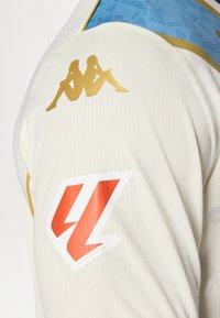 Krem jersej s plavim i zlatnim detaljima, izrađen od teksturiranog materijala. Prikazuje zlatni logo na ramenu i veliki crveno-bijeli grb.