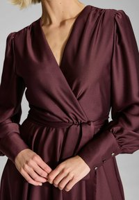 Robe portefeuille bordeaux en tissu lisse. Dotée de manches bouffantes, d'une ceinture à nouer à la taille et d'accents de boutons sur les poignets.