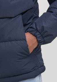 Mörkblå vadderad jacka med elastiska ärmslut, har en delvis öppen sidoficka, tillverkad av ett slätt, lätt material.