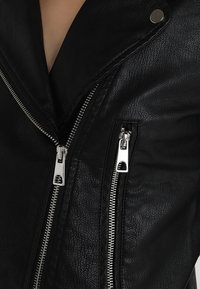 Veste en cuir noire avec une finition texturée, des fermetures éclair argentées proéminentes et un détail de bouton-pression sur un large col.