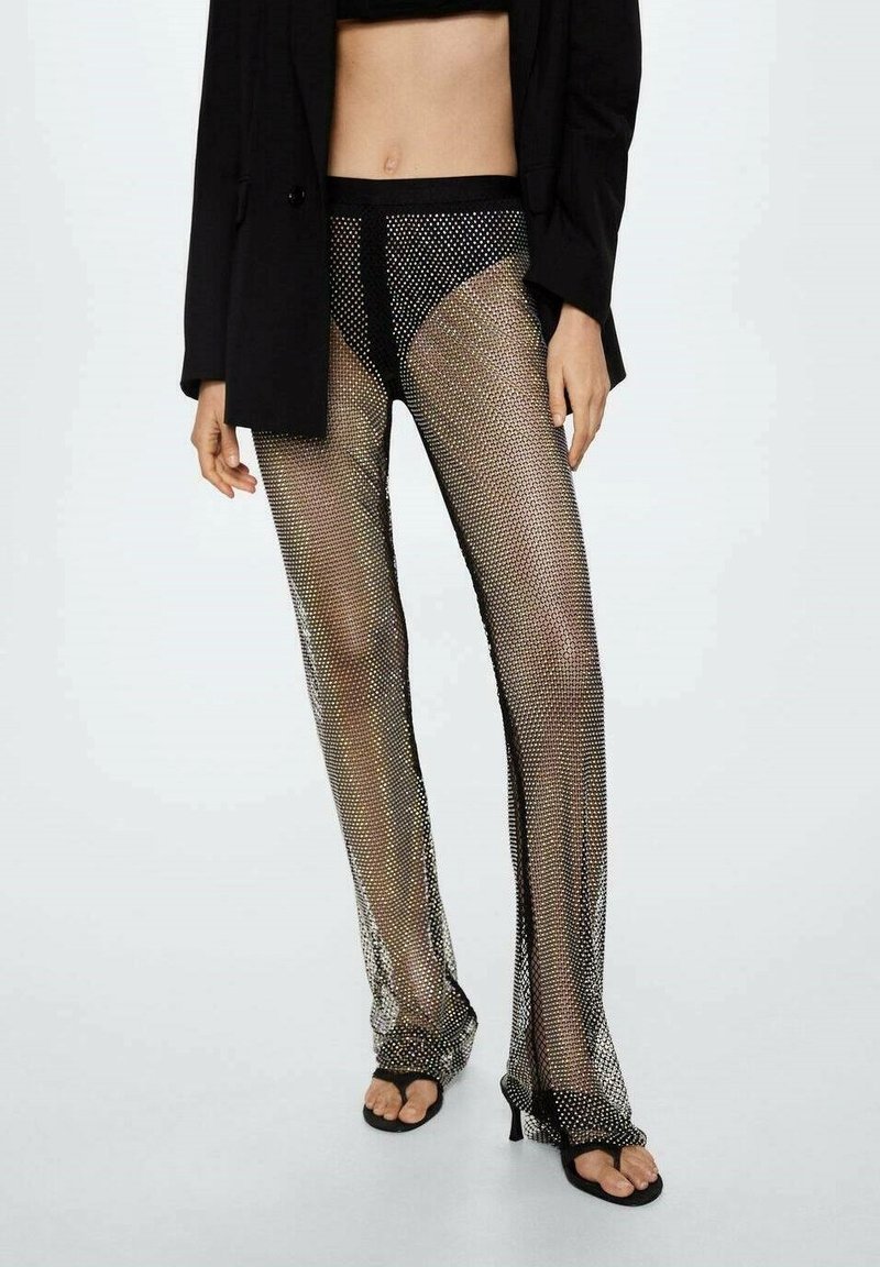 Femme portant un pantalon en résille noir avec sous-vêtements noirs, un blazer noir et des sandales noires à talons, debout devant un fond clair uni.