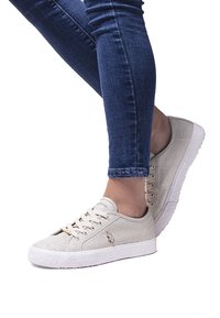 Feet wearing beige glittery U.S. Polo Assn. sneakers paired with blue skinny jeans against a white background.
