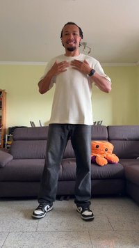 Homem de pé em ambiente interno, vestindo uma camisa branca e jeans pretos, sorrindo com as mãos levantadas ao peito, um polvo laranja de pelúcia no sofá atrás dele.