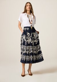 Cămașă albă cu mânecă scurtă, asortată cu o fustă maxi florală bleumarin, cu modele albe. Modelul poartă tocuri cu imprimeu de leopard și coliere suprapuse.