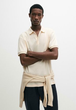 Giovane uomo nero con le braccia incrociate che indossa una polo color crema e un maglione chiaro legato in vita su uno sfondo bianco semplice.