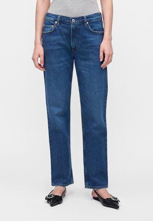 Person iført blå jeans med lige ben, en grå top og sorte spidse flade sko med spænde, stående mod en ensartet hvid baggrund.