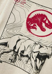 Camiseta gris con un diseño ilustrado de un T-Rex negro y un círculo de salpicadura roja, con líneas topográficas en el fondo.