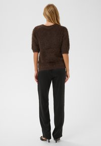 Femme aux longs cheveux blonds portant un pull court en laine marron, un pantalon noir droit et des chaussures noires à talons bas, debout de dos.