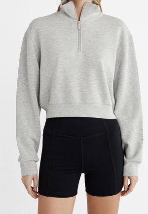 Person iført en lysegrå half-zip pullover sweatshirt og tætsiddende sorte højtaljede shorts mod en hvid baggrund.