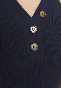 Prenda de punto acanalado en azul marino con escote en V, que presenta tres botones redondos en diferentes tonos de beige y marrón. Detalle de tela texturizada visible.