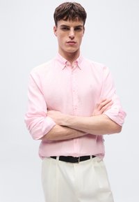 Νέος άνδρας με κοντά καστανά μαλλιά, που φοράει ένα ανοιχτό ροζ πουκάμισο με κουμπιά και γυρισμένα μανίκια και λευκό παντελόνι, με σταυρωμένα χέρια.