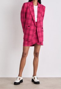 Růžový károvaný blazer s dvouřadovým zapínáním, doplněný o sladěnou mini sukni. Modelka nosí bílý top a korpulentní bílé boty s černými podrážkami.