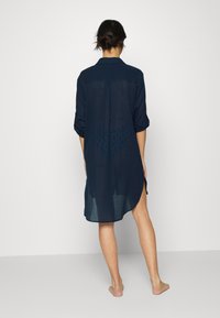 Navy blue shirt dress, lightweight fabric, long sleeves rolled up, button-up design, side slits, sheer back with geometric pattern.