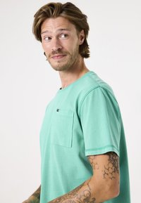 Groene katoenen T-shirt met een ronde hals, korte mouwen en een linker borstzak. Opvallende contrasterende stiksels langs de naden.