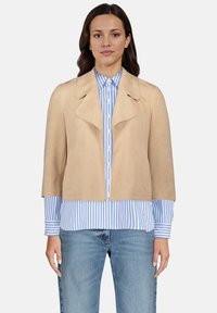 Veste en suède beige avec un large col et des manches courtes, portée sur une chemise à rayures bleu et blanc avec boutons, assortie à un jean clair.