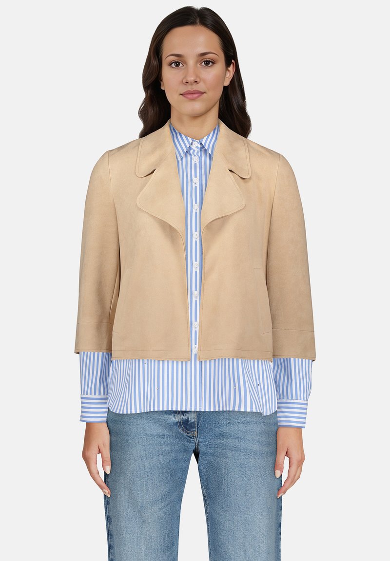 Veste en suède beige avec un large col et des manches courtes, portée sur une chemise à rayures bleu et blanc avec boutons, assortie à un jean clair.