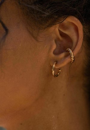 Boucles d'oreilles en or en forme de créole présentant des textures torsadées et lisses, mettant en avant une finition brillante avec un design minimaliste, portées à l'oreille.