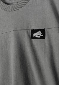 Plan rapproché d'une chemise en tissu gris montrant un col côtelé et un patch rectangulaire noir avec le texte stylisé blanc « unity » sur la poitrine.