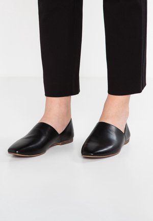Persoon die zwarte cropped broek en zwarte leren puntige instapschoenen draagt, staande op een witte vloer.