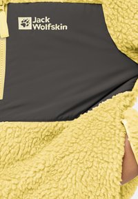 Keltainen fleece-takki, jossa on sileä harmaa paneeli, varustettu vetoketjulla ja vaaleanharmaalla Jack Wolfskin -logolla. Teksturoitu pinta ja lämmin muotoilu.