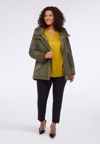 Olivgrüne gepolsterte Jacke mit abnehmbarer Kapuze, Reißverschlusstaschen und silbernem Zubehör, getragen über einer gelben Bluse und schwarzen Hosen.