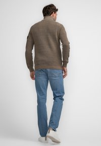 Suéter de punto marrón con cuello alto, combinado con jeans de color azul claro y zapatillas blancas. El suéter presenta una textura acanalada y un corte ajustado.