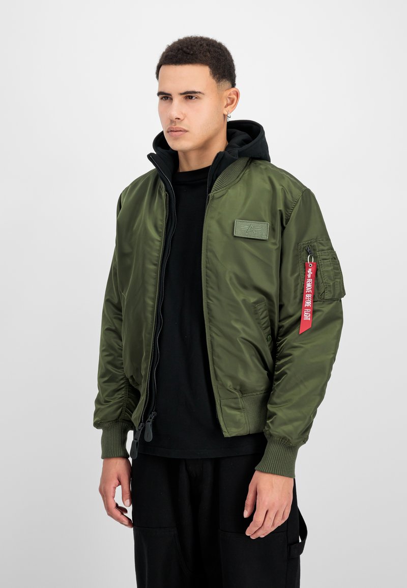 Junger Mann trägt grüne Bomberjacke mit rotem „Remove Before Flight“-Anhänger am Ärmel, schwarzen Hoodie und schwarze Hose, blickt nach links.
