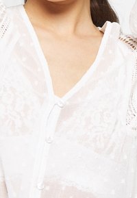 Weiße Spitzenbluse mit V-Ausschnitt, Knopfverschluss, strukturierten Polka-Dots und transparentem Stoff, mit aufwendigen Spitzen-Details an den Schultern.