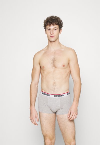Shirtless man with curly hair wearing gray Moschino boxer briefs standing against a plain white background.