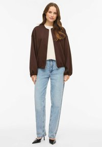 Pruun lukuga bomber-jakk õhupallivarrukatega, kombineeritud helesiniste sirgete teksade ja mustade teravate kontsakingadega. All on valge topp.