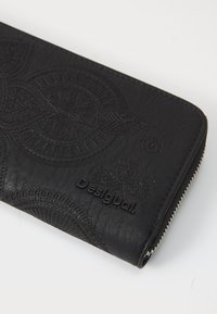 Portafoglio in pelle nera con motivi in rilievo, superficie testurizzata e chiusura con zip, con un logo che recita "Desigual."