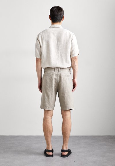 Chemise en lin à manches courtes et shorts beiges. Conception ajustée, texture unie et poches au dos. Associée à des sandales noires.