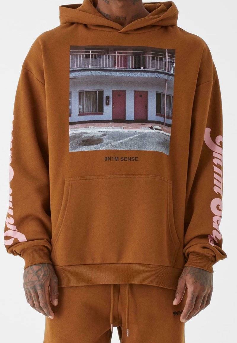 Personne portant un sweat à capuche marron avec une photo en façade d'un bâtiment avec deux portes roses et le texte "9N1M SENSE." sur la poitrine ainsi qu'un texte rose sur les manches.