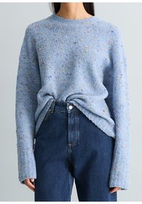 Personne portant un pull tricoté texturé bleu clair avec des taches multicolores, rentré dans un jean taille haute bleu foncé sur un fond uni.