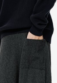 Kéz a sötétszürke texturált nadrág zsebében, látható a sötétkék kötött pulóver mandzsettája.