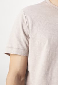Gros plan sur un t-shirt à manches courtes beige clair sur le bras supérieur et l'épaule d'une personne, sur un fond blanc.