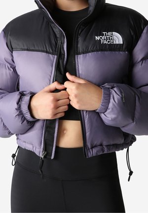 Osoba ubrana w fioletowo-czarną puchową kurtkę North Face, częściowo rozpiętą, odsłaniającą krótki czarny top i czarne legginsy.