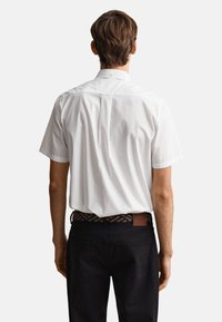 Man standing facing away, wearing a white short-sleeve button-up shirt and black pants with a patterned belt against a plain background.