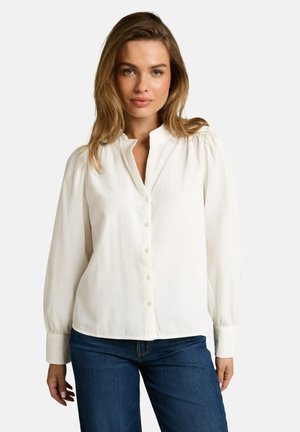 Vrouw met lichtbruin haar die een witte blouse met knopen en blauwe spijkerbroek draagt, staand tegen een effen achtergrond.