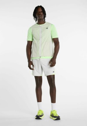 Mann steht und trägt ein hellgrünes kurzärmeliges Hemd, beige ärmellose Weste, weiße Shorts, weiße Socken und neongelbe-schwarze Laufschuhe.