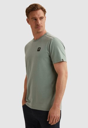 Man draagt een lichtgroene T-shirt met korte mouwen en een zwart logo op borst en mouw, kijkt naar links met handen in zijn zakken.