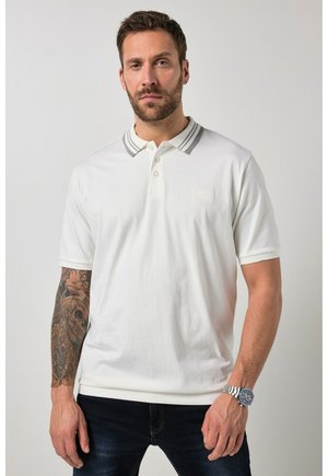 Mann mit Bart, der ein weißes Poloshirt mit grauem gestreiftem Kragen, dunkle Jeans, eine Armbanduhr und ein detailliertes Totenkopf-Tattoo am linken Unterarm trägt.