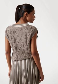 Knit sweater in taupe with intricate cable patterns, short sleeves, and ribbed neckline. Paired with a pleated, stripe-patterned skirt.