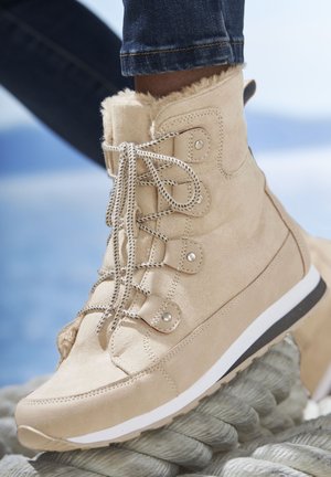 Beige Wildleder-Stiefeletten mit Fellbesatz, runder Zehenpartie, Schnürdesign, metallischen Ösen und weißer Mittelsohle auf einer strukturierten Sohle.