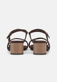 Ζευγάρι σκούρα καφέ σανδάλια με λουράκια και χαμηλά τετράγωνα τακούνια, απεικονιζόμενα από πίσω σε λευκό φόντο.