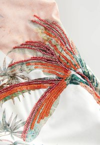 Broderat tyg med orange och röda paljetter på en ljus bakgrund, med ett mönster av tropiska blad och en mjuk, texturerad yta.