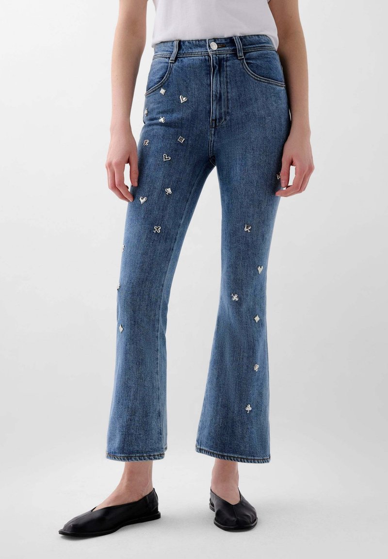 Jean en denim à jambes évasées avec de petits symboles métalliques de jeux de cartes dispersés à l'avant, porté avec des ballerines noires et un haut blanc.