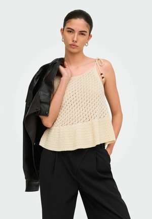 Mujer joven que lleva un top de punto sin mangas beige con volantes, pantalones negros y sostiene una chaqueta de cuero negra sobre su hombro.