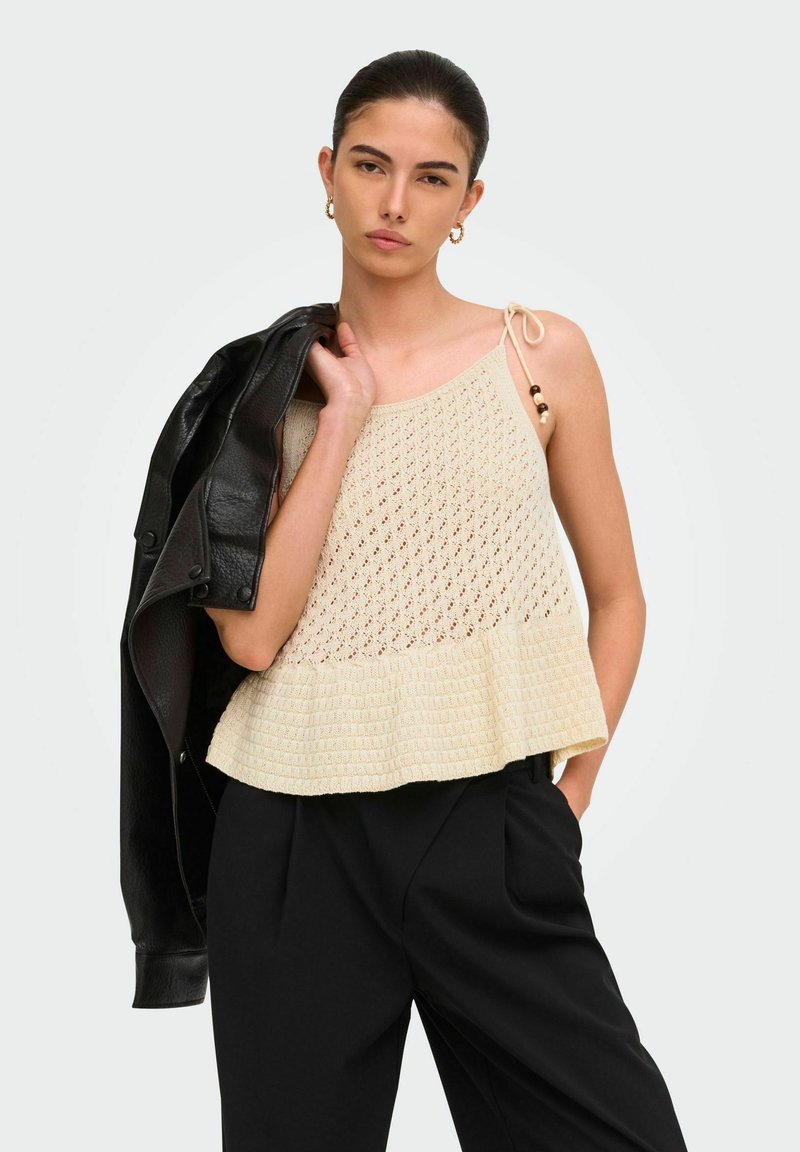 Jeune femme portant un haut en tricot beige sans manches avec des volants, un pantalon noir, et tenant une veste en cuir noire sur son épaule.